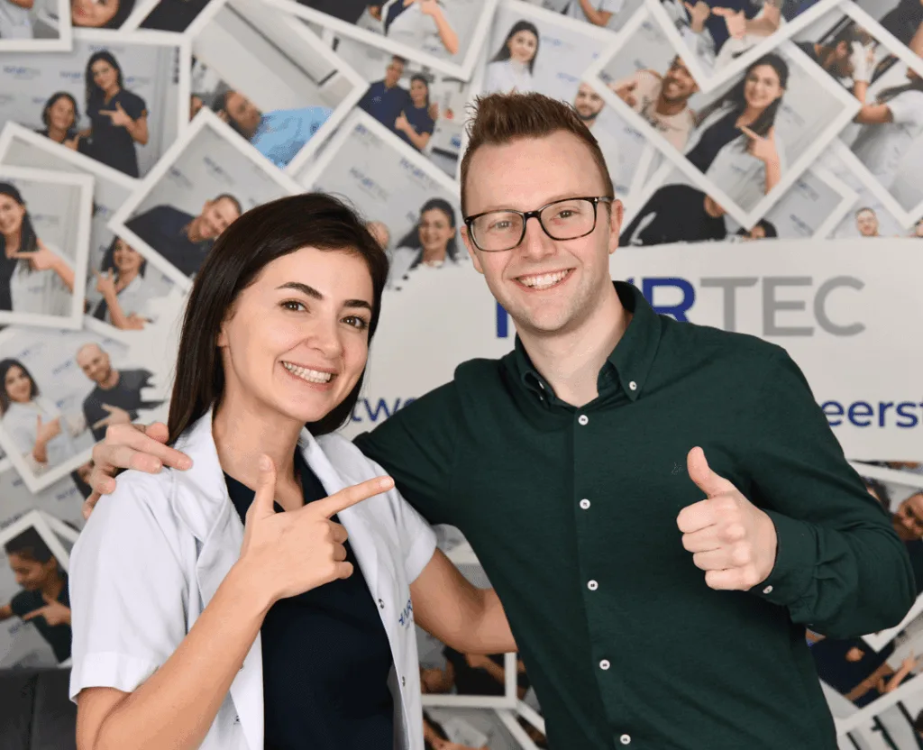 Medewerker van Hairtec Haarkliniek in witte jas wijst lachend naar een man die zijn duim opsteekt, voor een wand vol foto's